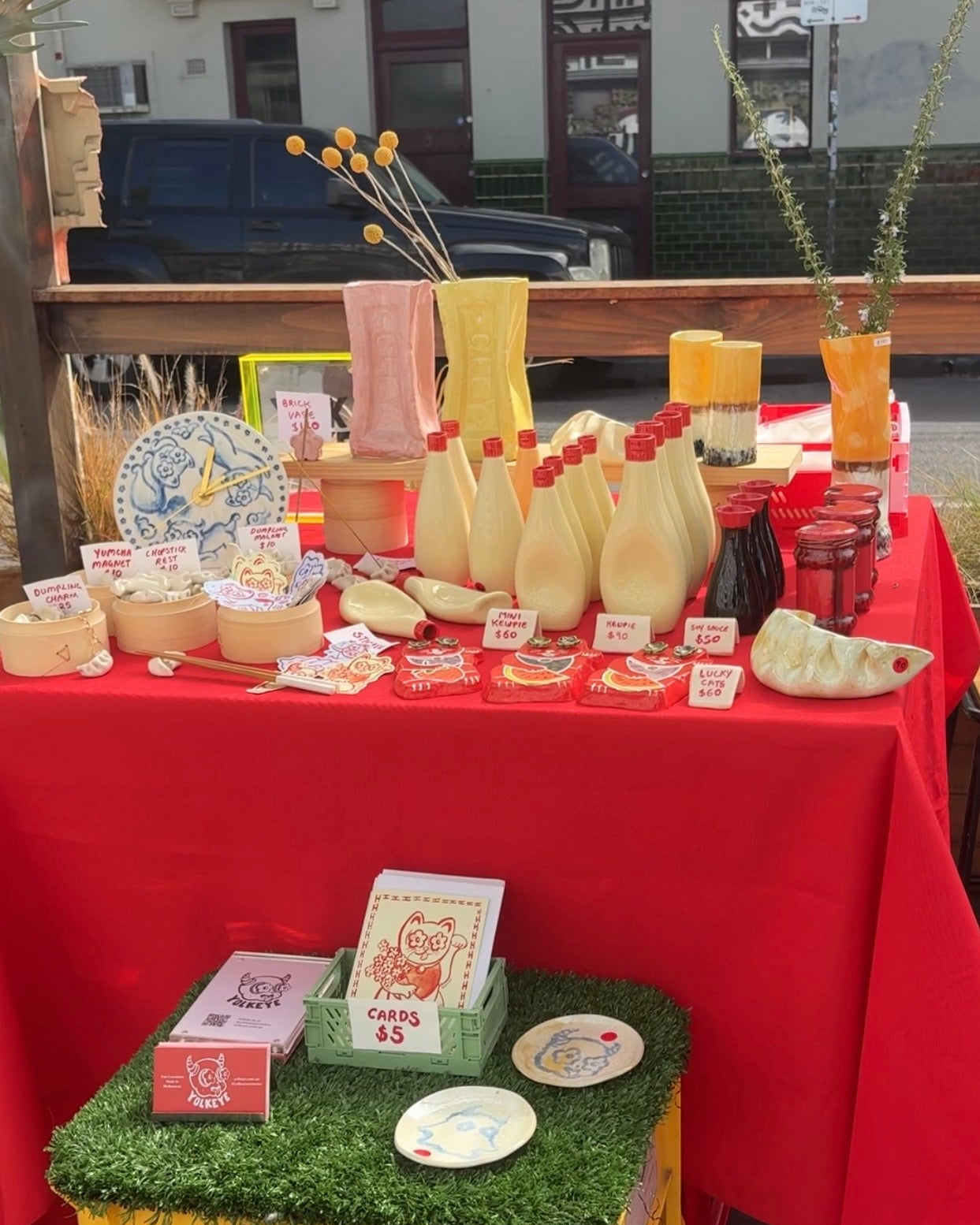 A market stall table covered with a bright red tablecloth, displaying handmade crafts and small products including cream-colored bottles, decorative plates, small boxes, and vases. A yellow crate sits in front on artificial green grass.